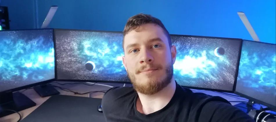 A man with a beard and short hair takes a selfie in front of a multi-monitor gaming setup. The monitors display a blue nebula background with a crescent moon. Two light bars are visible above the monitors.