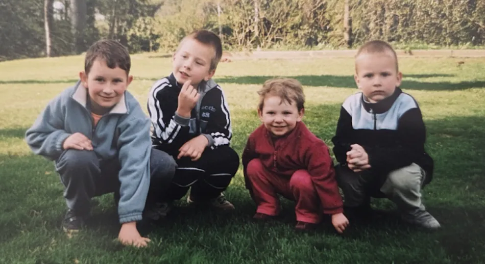 Four young boys are crouched on a grassy lawn, looking towards the camera. The boy on the far left wears a blue jacket and smiles, the boy next to him in a black and white track suit smiles while looking up and to the left, the third boy in a red fleece smiles, and the boy on the far right in a black and light blue sweater has a serious expression.
