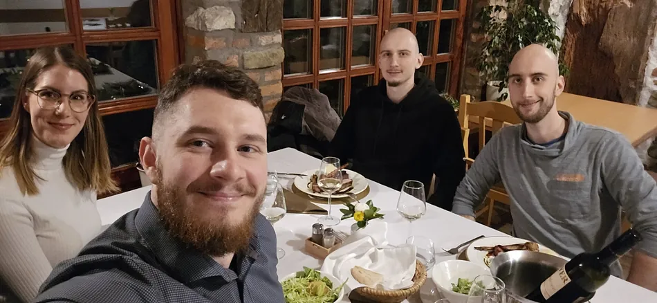 A group of four people, two men and two women, are seated around a white table at a restaurant, smiling at the camera. Food and wine glasses are visible on the table.